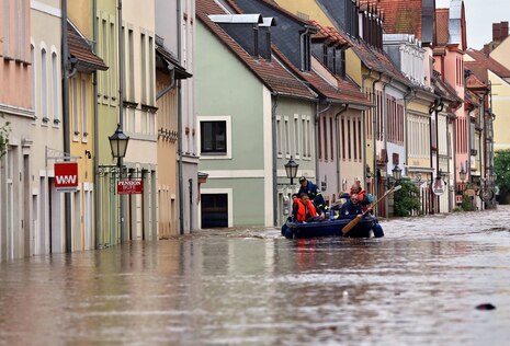 Bild einer überfluteten Innenstadt, mittig fährt ein Rettungsboot