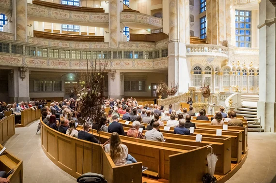 Menschen sitzen in einer Kirche. An einem Rednerpult vor ihnen steht eine Frau,