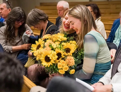Zwei Frauen umarmen sich. Eine von ihnen hält einen Blumenstrauß in der Hand.